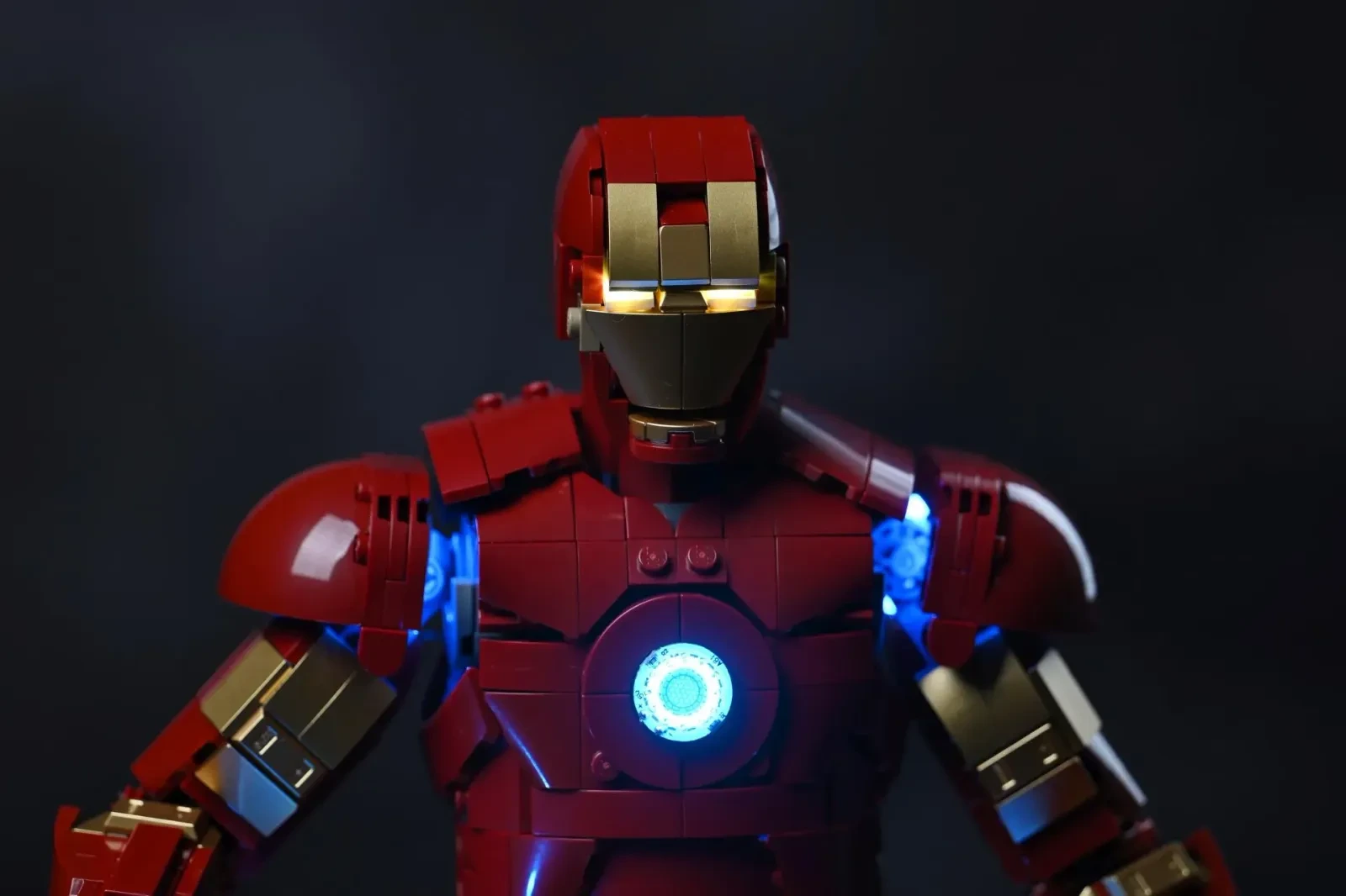 Der Oberkörper der LEGO-Iron-Man-Mark-3-Figur in einem dunklen Raum, Brustreaktor und Schulter-Repulsoren leuchten blau, die Augen golden
