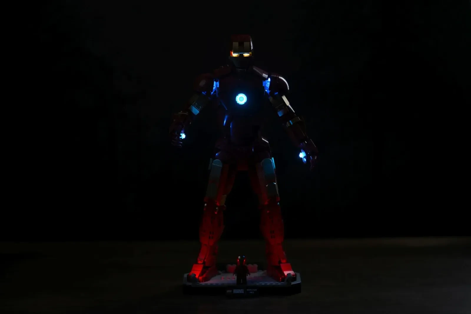 Die LEGO-Iron-Man-Mark-3-Figur in fast völliger Dunkelheit mit eingeschaltetem BriksMax-Kit, Brustreaktor und Repulsoren leuchten, rotes Unterglühen erhellt die Base