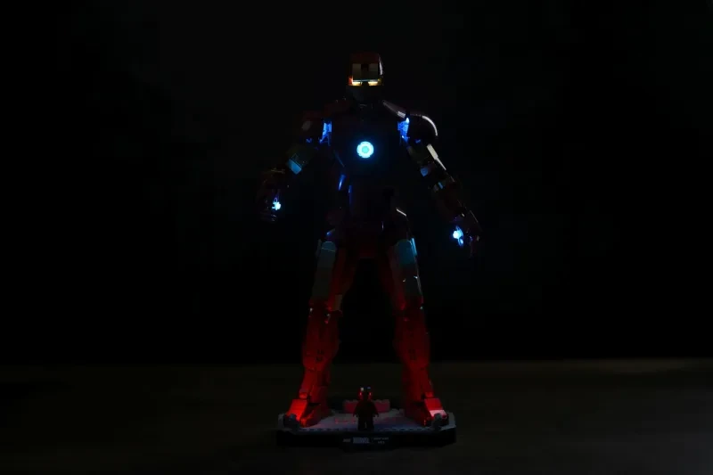 Die LEGO Iron Man Mark 3 Figur in fast völliger Dunkelheit, Brustreaktor und Repulsoren leuchten, rotes Unterglühen erhellt die Display-Base
